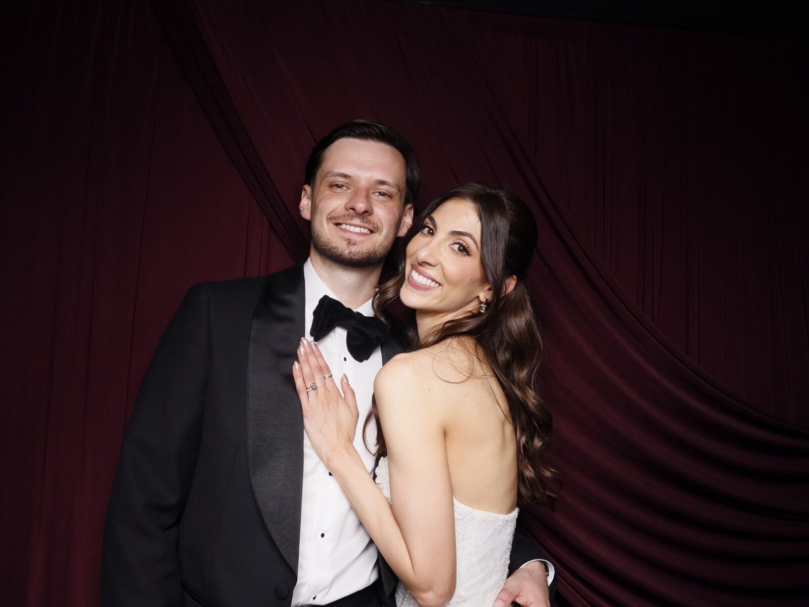 Bride with her hand resting on the groom's chest showing her rings, both smiling at the camera in front of a deep burgundy draped backdrop