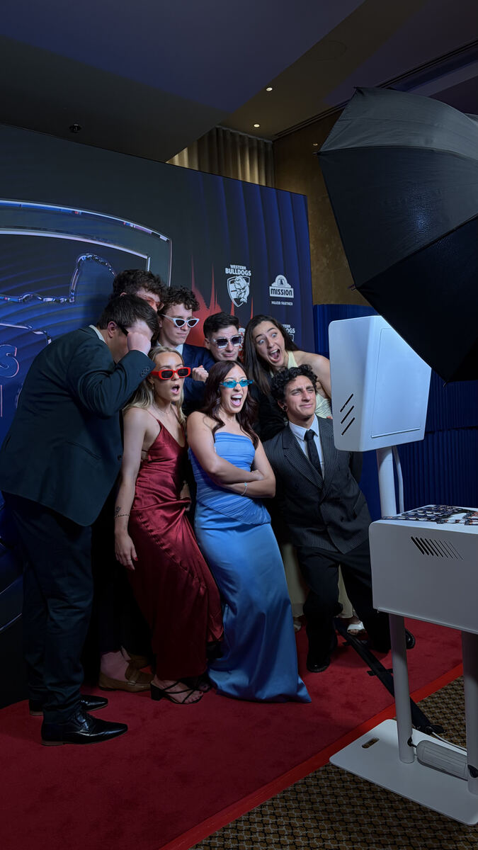 Group of guests in formal wear and novelty sunglasses posing on a red carpet in front of a Western Bulldogs and Mission backdrop, with a white photo booth and black umbrella light on the right