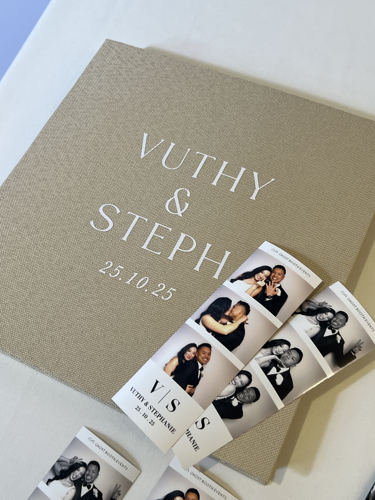 A sand-coloured linen guestbook printed with Vuthy and Stephanie and the date, with black and white photo strips of the couple laid across it