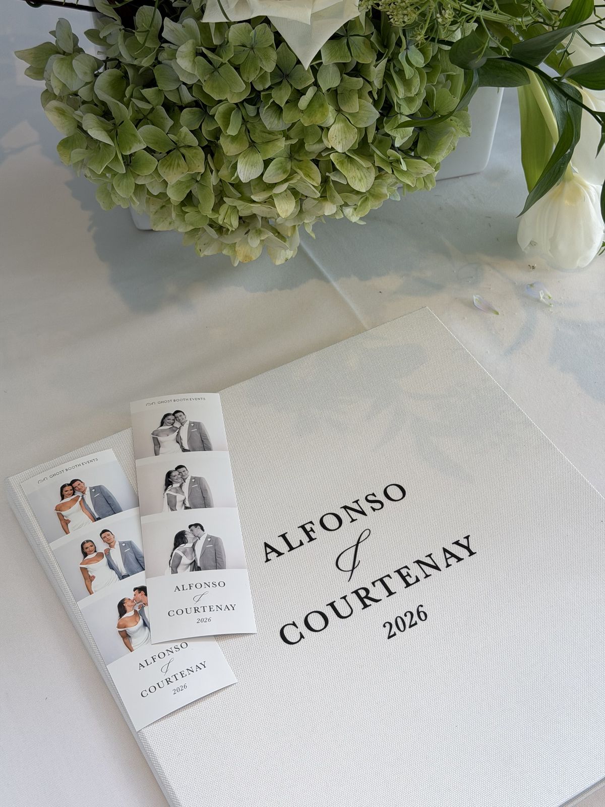 A white linen guestbook printed with Alfonso and Courtenay 2026, with two black and white photo strips of the couple resting on the cover