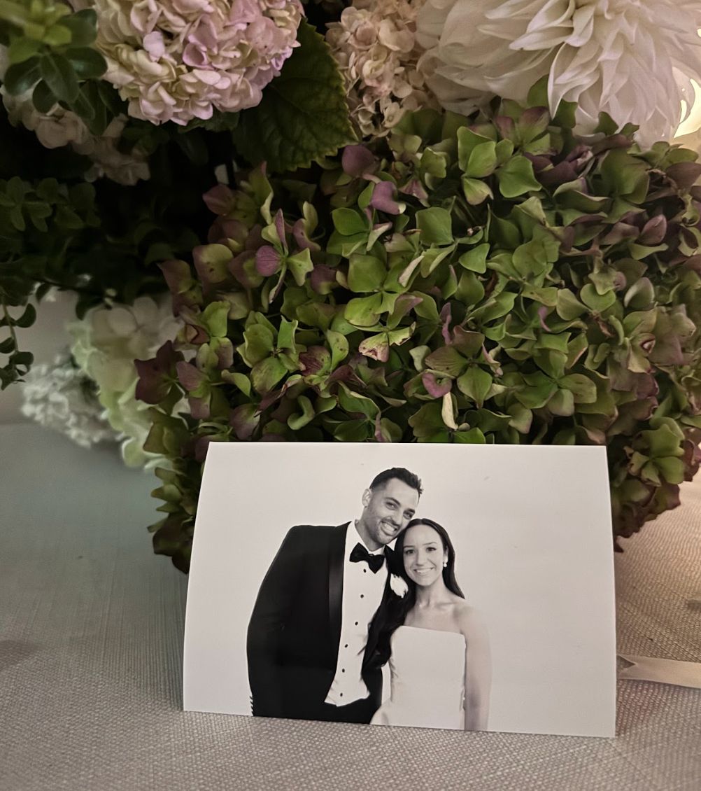 A black and white photo booth print of a smiling bride and groom propped against hydrangeas on a reception table