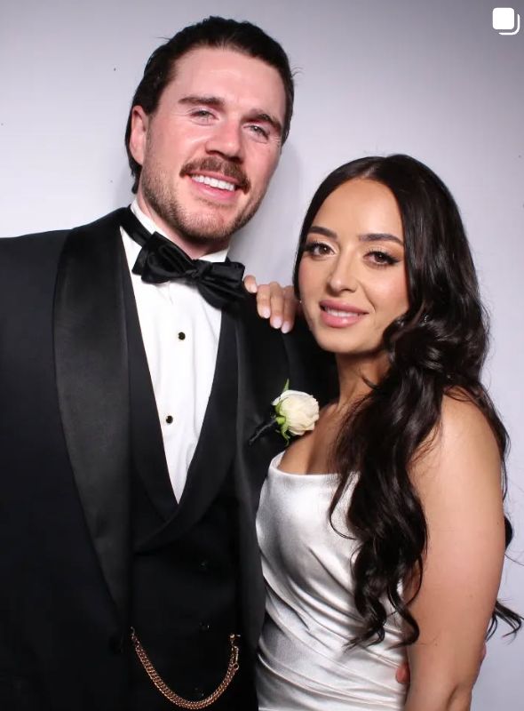A couple in black tie and a satin dress smiling together in front of a white photo booth backdrop