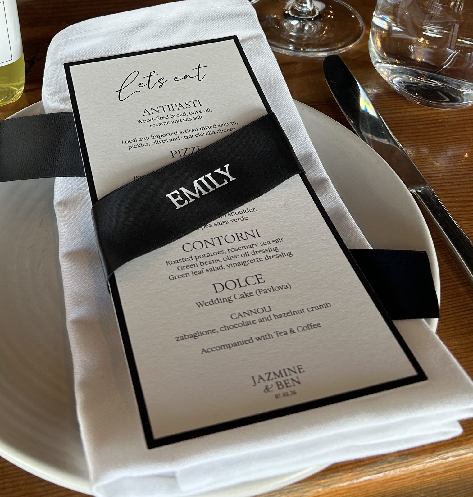 Printed wedding menu reading Let's eat with a black satin ribbon and silver name EMILY, on a folded white napkin at a place setting