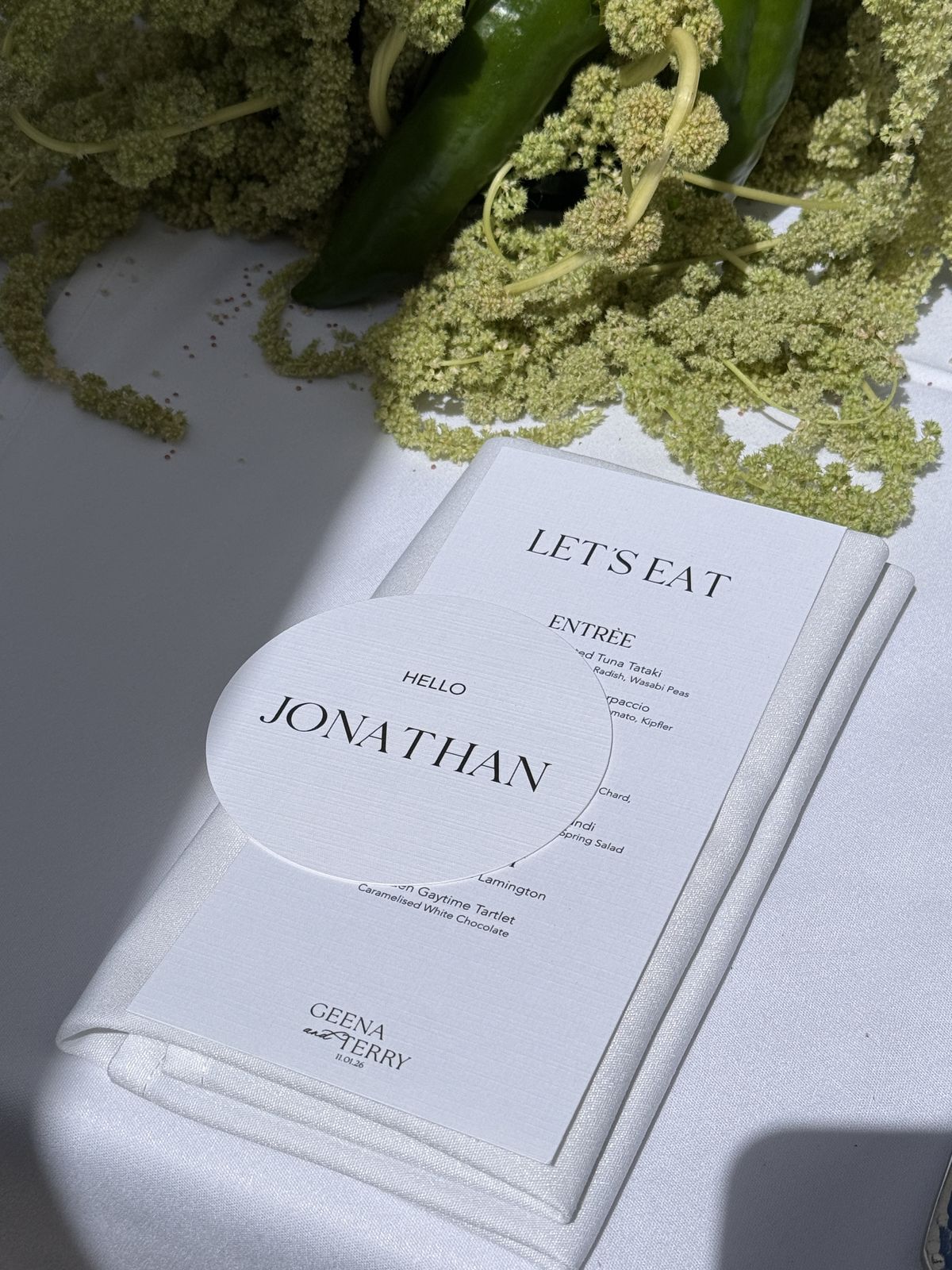 Wedding place setting with a Let's Eat menu and a round Hello Jonathan name card beneath green floral styling