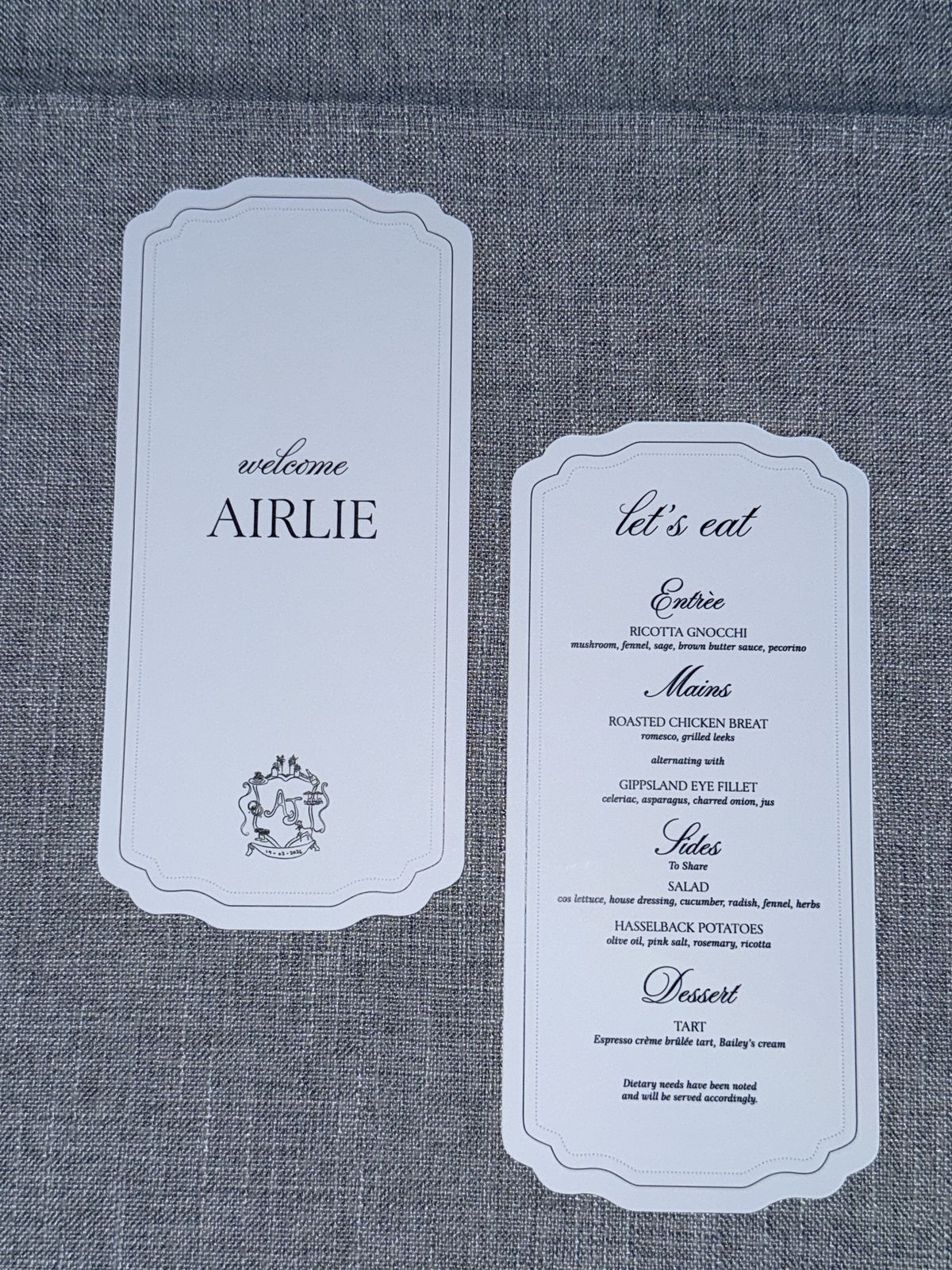 Welcome name card and Let's Eat menu with entree, mains, sides and dessert, laid on grey linen