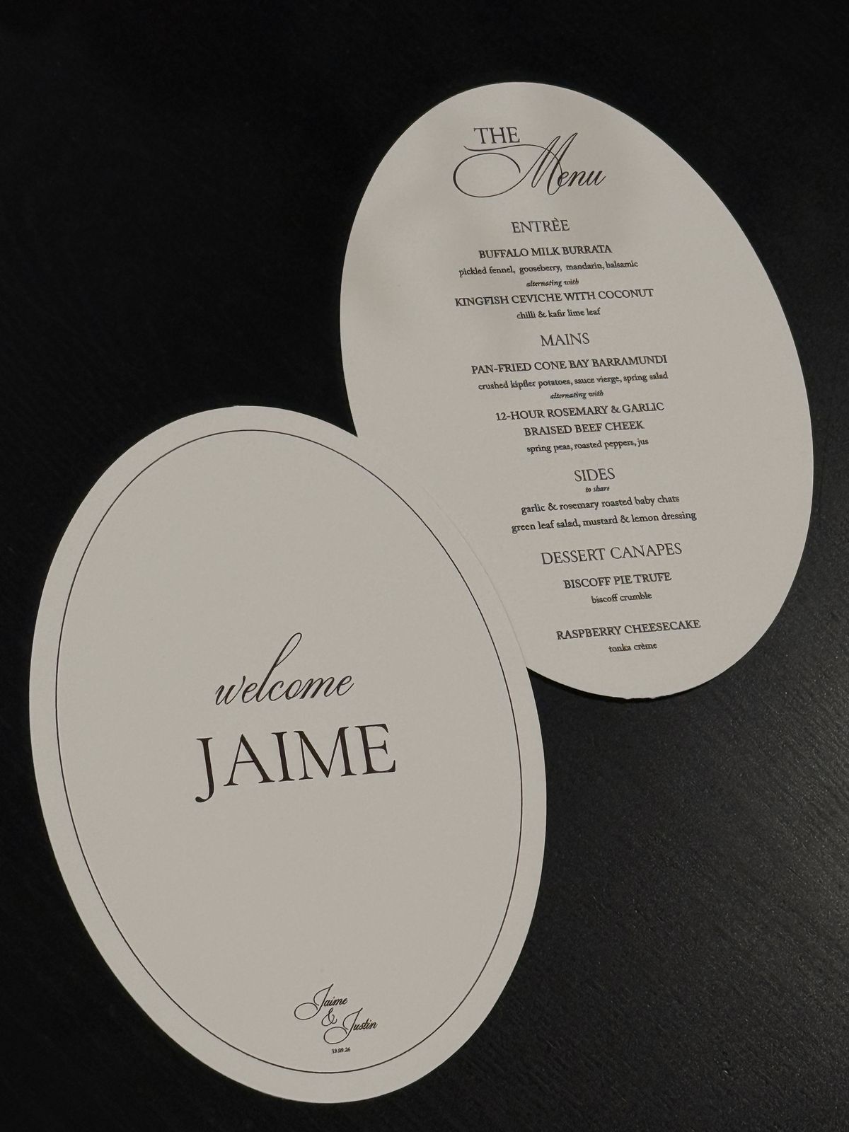 Oval Welcome Jaime place card beside an oval wedding menu on a black table