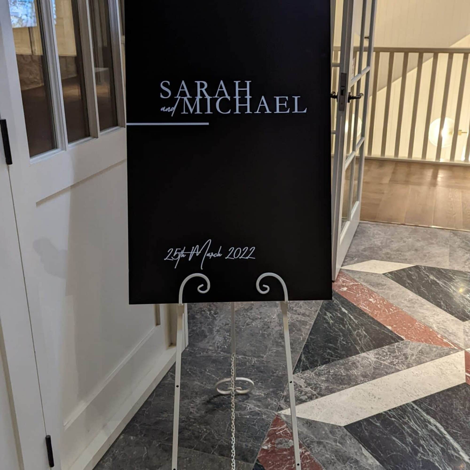 Black acrylic wedding welcome sign with white lettering for Sarah and Michael on a white metal easel with scrolled feet