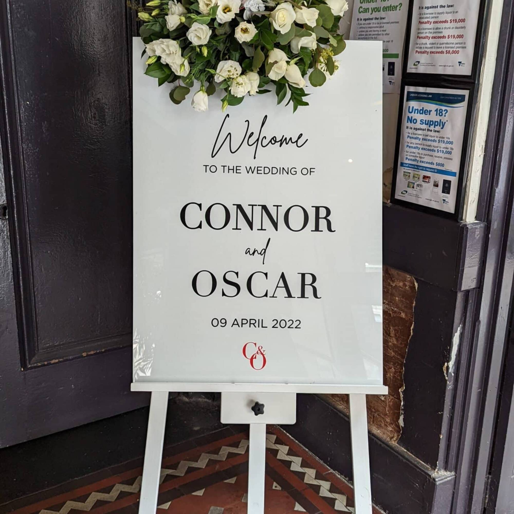 White wedding welcome sign reading Welcome to the wedding of Connor and Oscar, 09 April 2022, on a white timber easel with white flowers on top