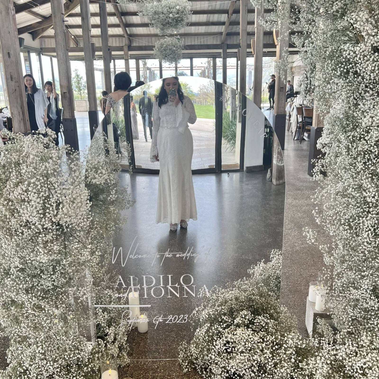 Mirror arch welcome sign framed by baby's breath, clearly reflecting a guest in a white outfit taking a photo of it