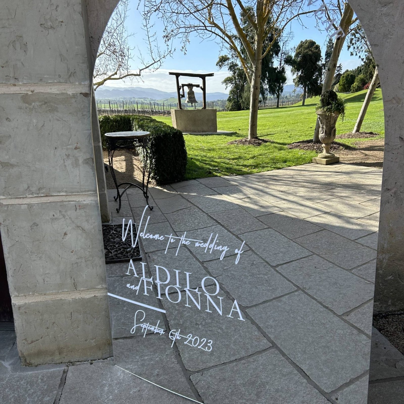Mirror wedding welcome sign under a sandstone archway looking out over a sunny lawn and garden