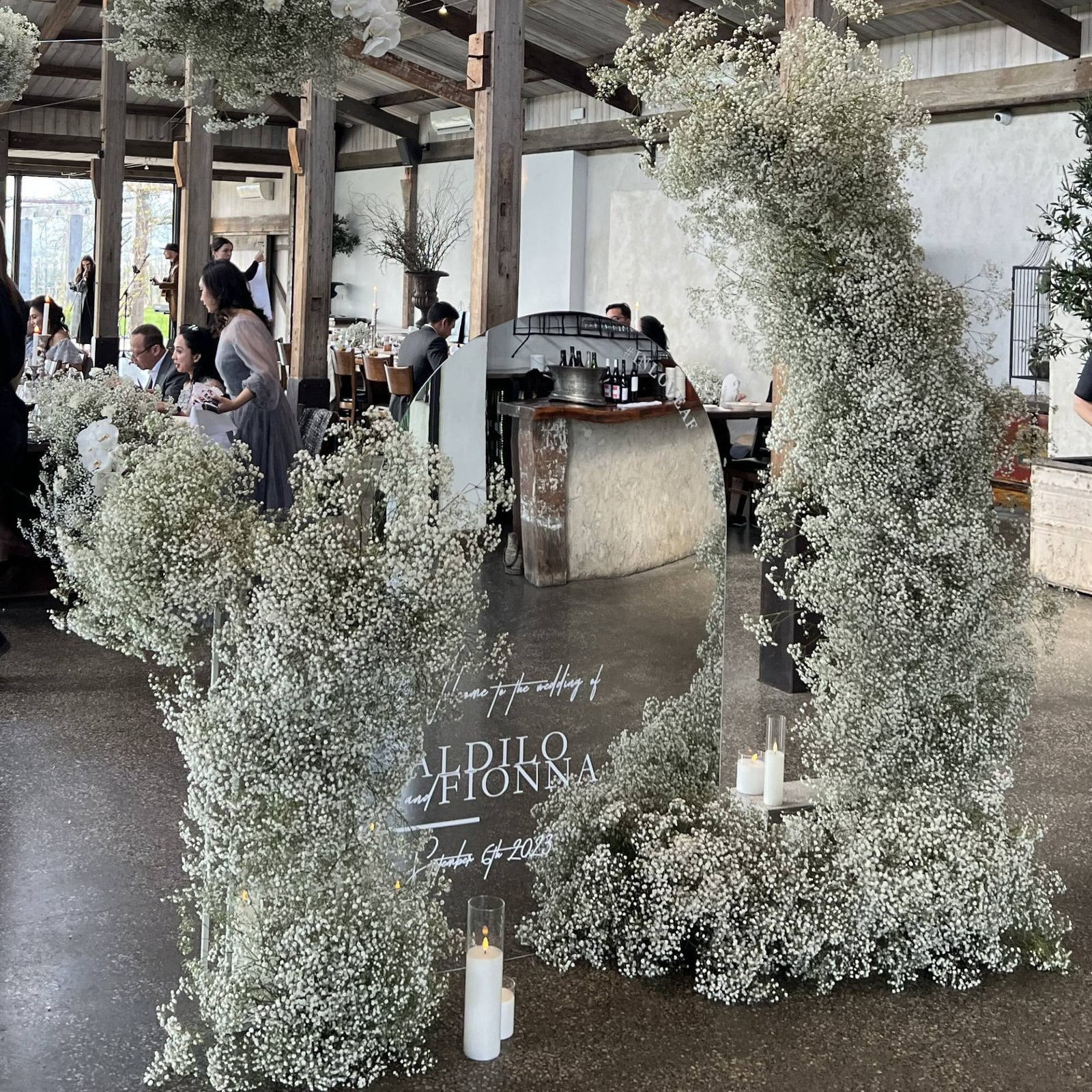 Mirror welcome sign surrounded by a tall, dense arch of baby's breath inside a timber-beamed reception venue