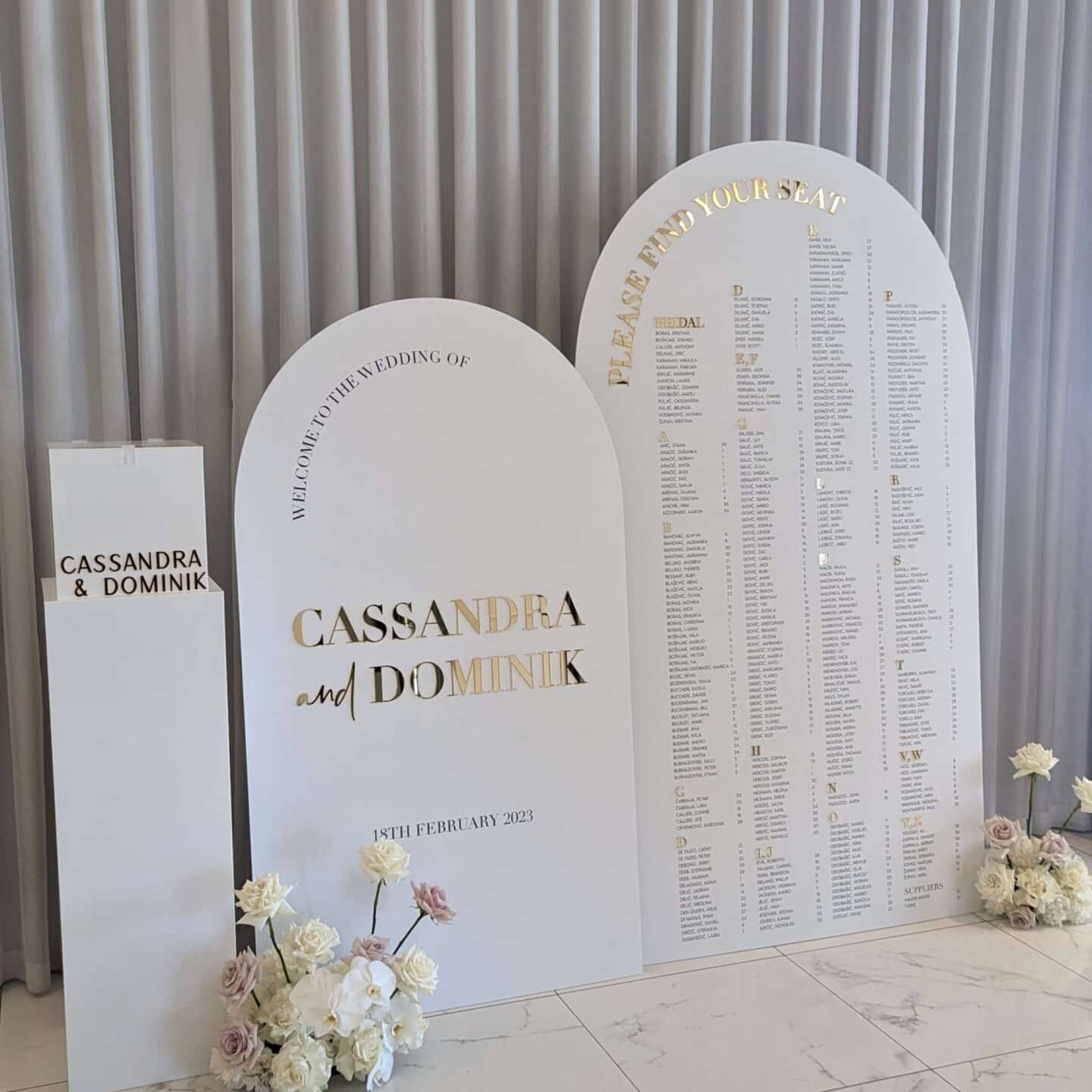 White arch welcome sign with gold mirror lettering for Cassandra and Dominik beside a white arch seating chart and a small white plinth