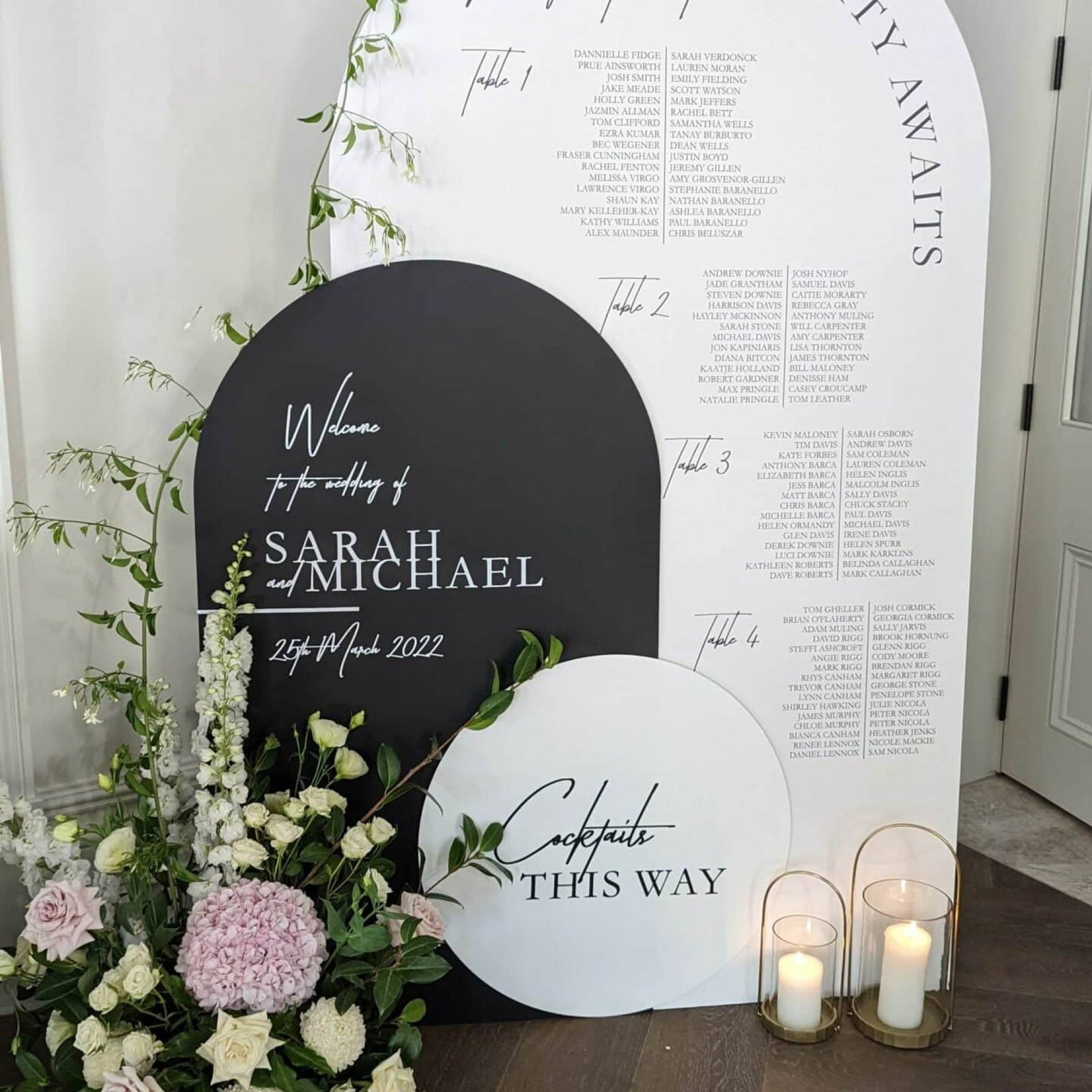 White arch seating chart with guests grouped under Table 1, Table 2, Table 3 and Table 4, behind a black arch welcome sign for Sarah and Michael and a white round sign reading Cocktails this way