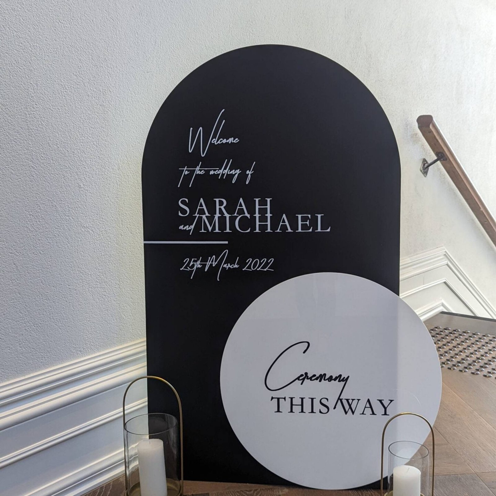 Black arched wedding welcome sign beside a round white Ceremony This Way sign at the foot of a staircase