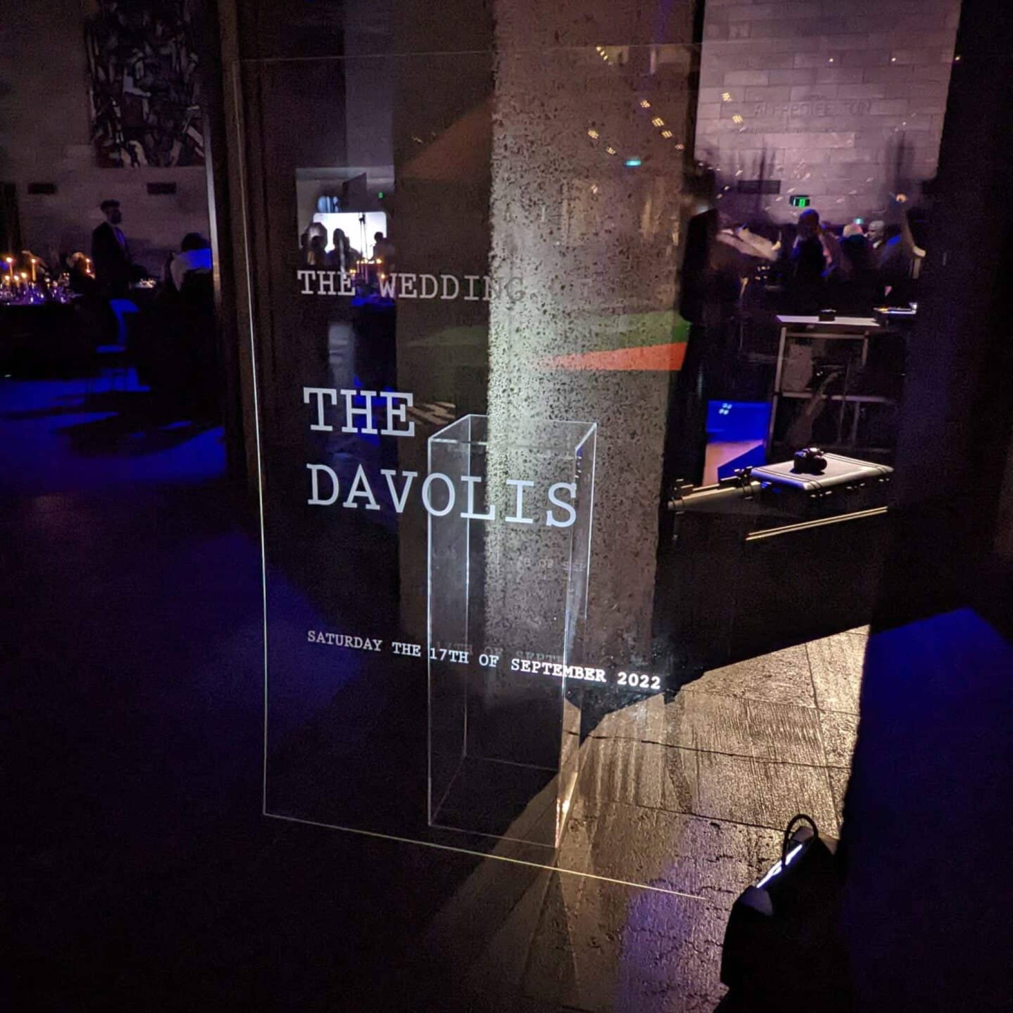 Clear acrylic welcome sign reading The wedding of The Davolis, Saturday the 17th of September 2022, glowing in a dark reception room with blue lighting