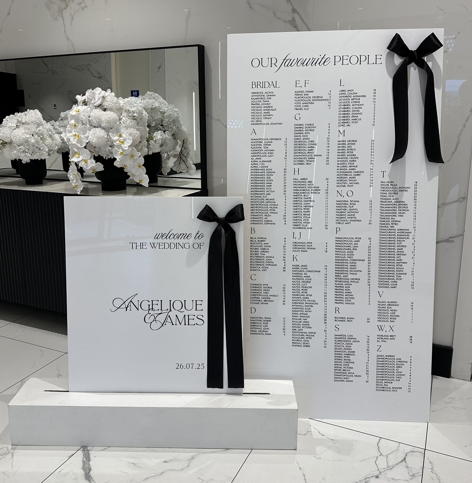 White acrylic welcome sign with a large black velvet bow, standing on a white plinth in front of a matching seating chart that also has a black bow
