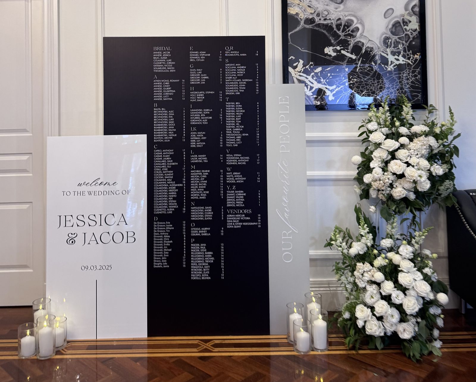 Black seating chart with white lettering behind a white welcome sign for Jessica and Jacob, with a narrow grey panel reading Our favourite people vertically, white florals and candles