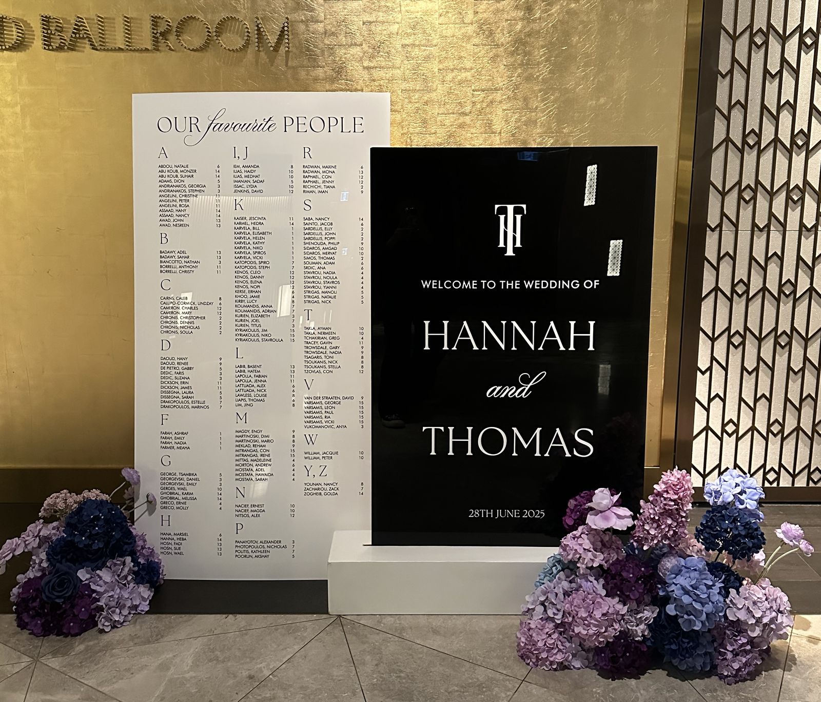 Black acrylic welcome sign with a white monogram above the names Hannah and Thomas, beside a white seating chart, with purple and blue hydrangeas at the base