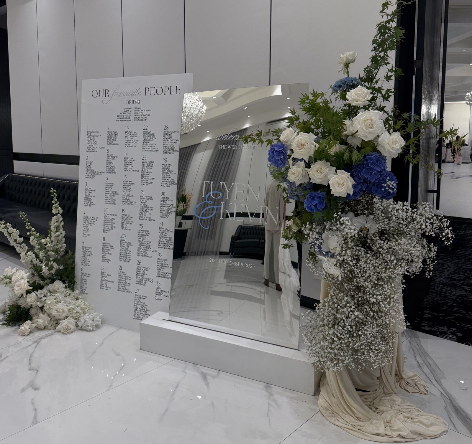 White acrylic seating chart headed Our favourite people with guests grouped under numbered tables, beside a mirror welcome sign on a white plinth with blue hydrangeas and white roses