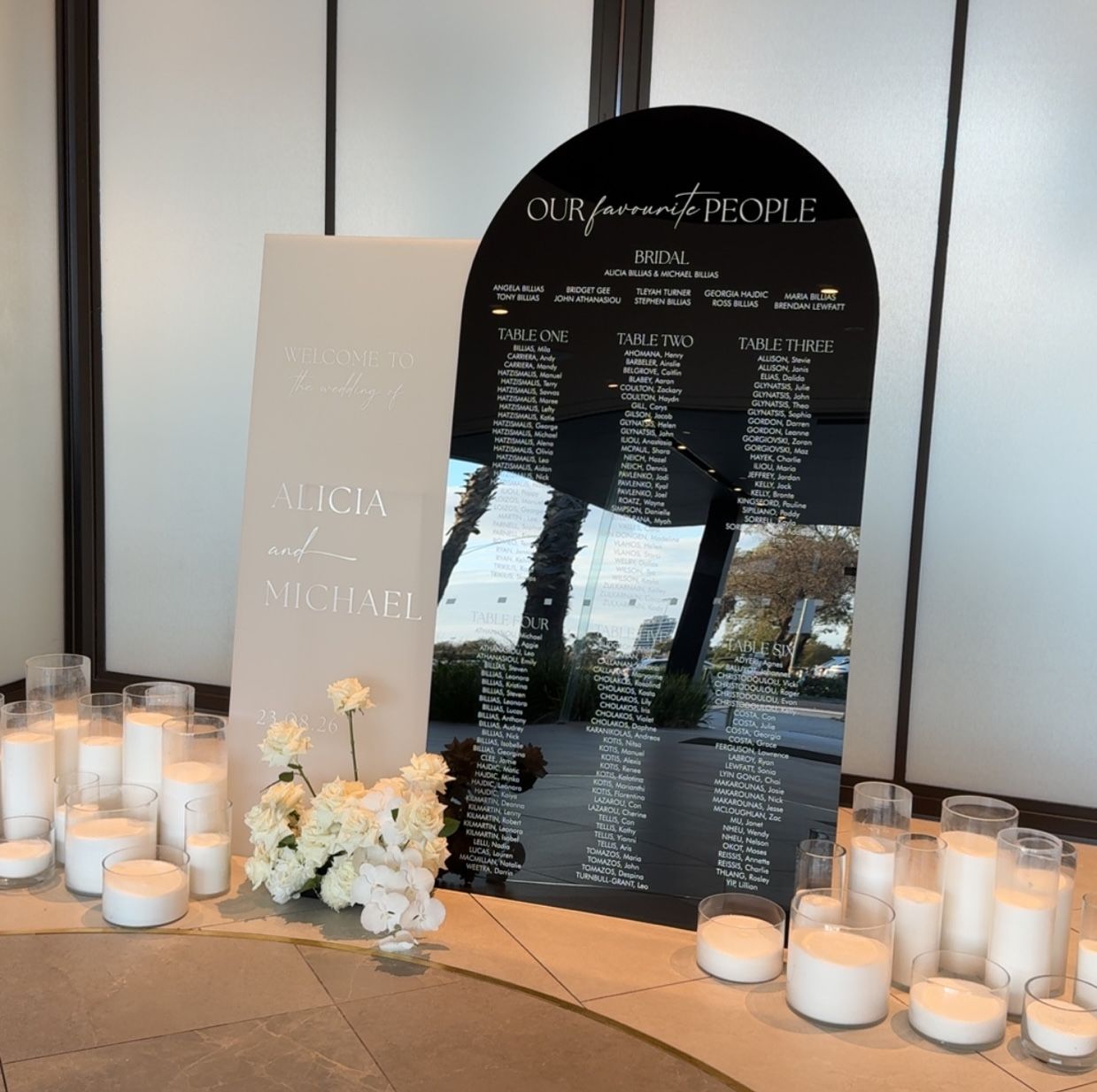 Black acrylic arch seating chart listing the bridal party then Table One, Table Two and Table Three, beside a cream welcome panel for Alicia and Michael, with candles in sand-filled glass
