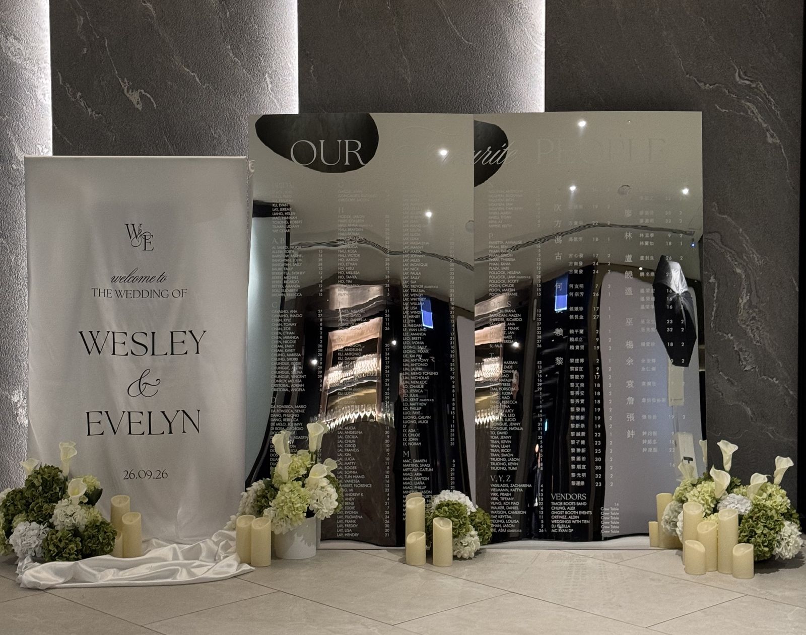 Two mirror seating chart panels, one listing guests in English and one in Chinese characters, beside a white fabric welcome sign with candles and white and green florals