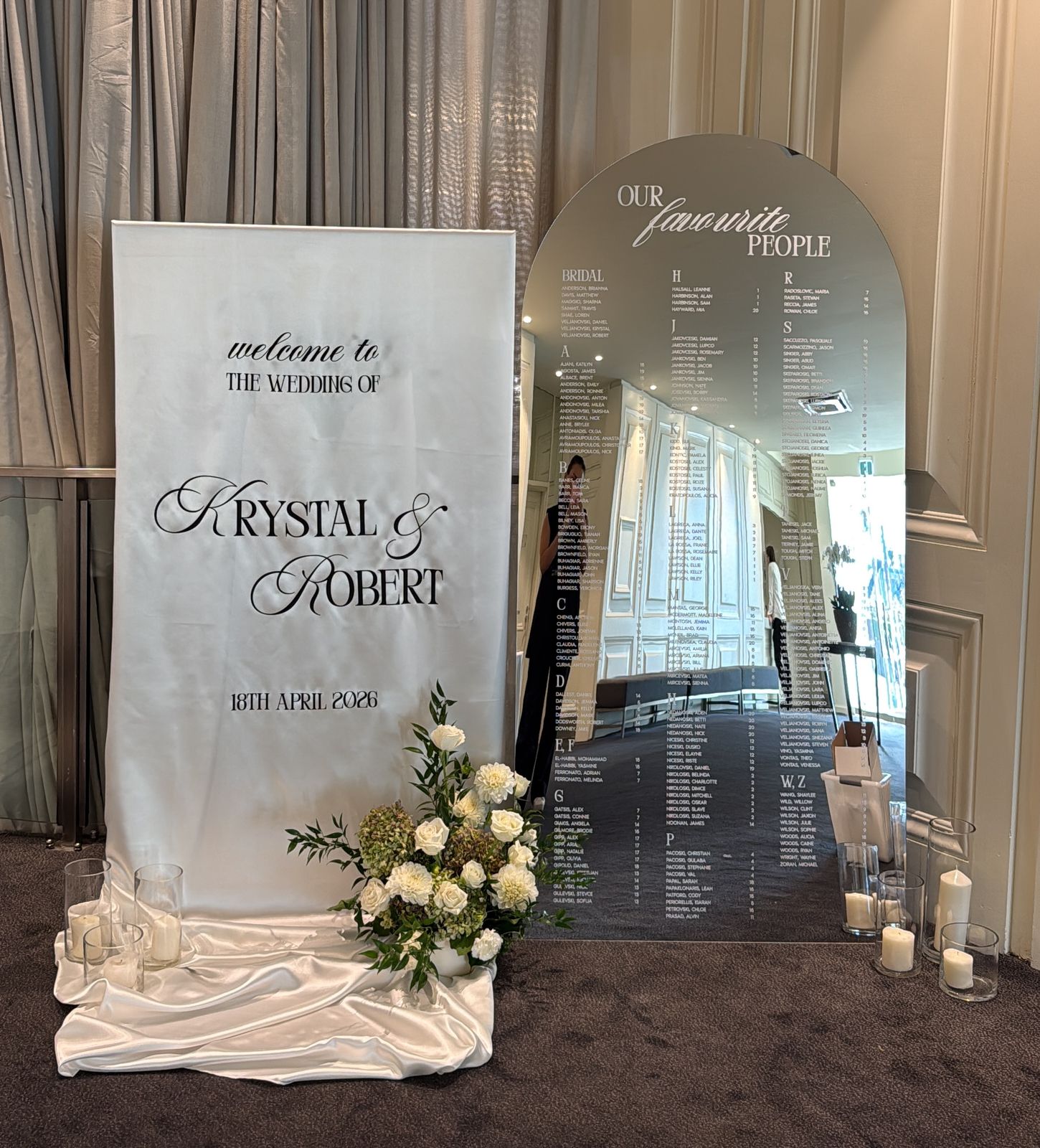 White fabric welcome sign reading welcome to the wedding of Krystal and Robert, 18th April 2026, beside a mirror arch seating chart, with white flowers and candles