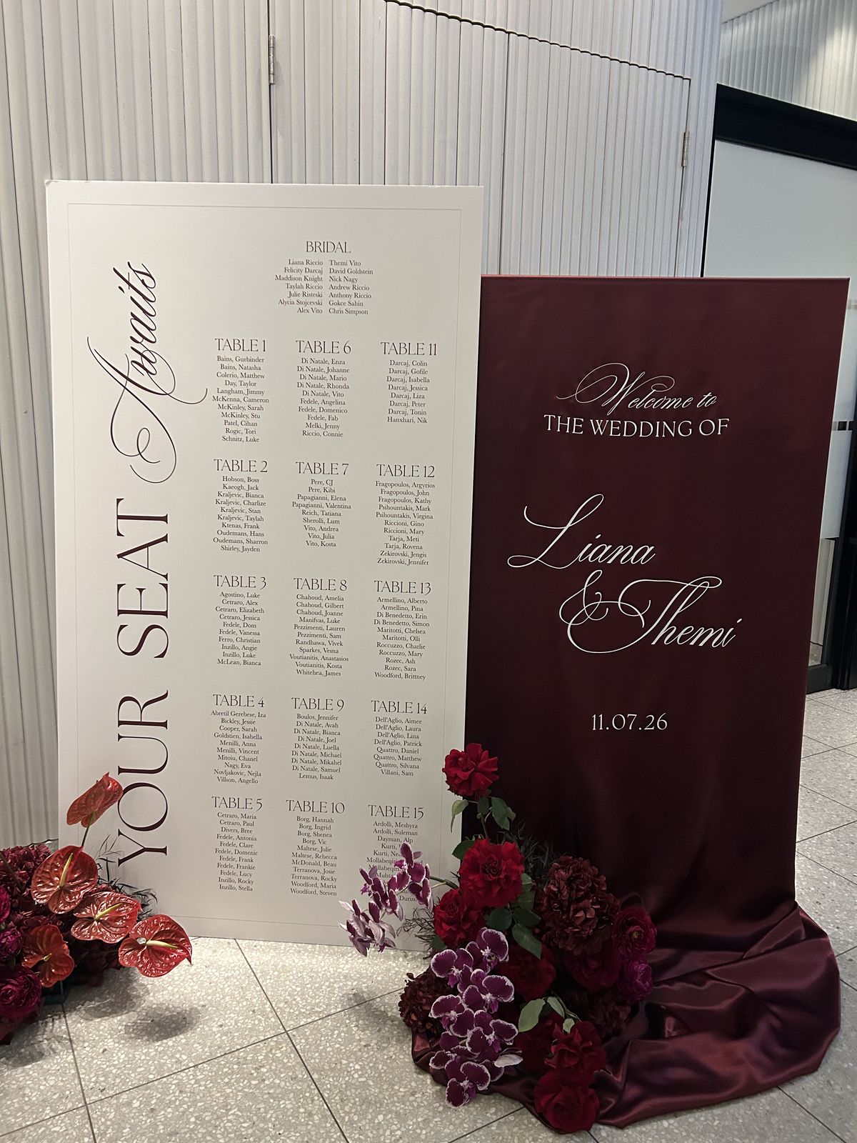 White seating chart with Your seat awaits written vertically and guests listed under Table 1 to Table 15, beside a burgundy satin welcome sign for Liana and Themi with red florals
