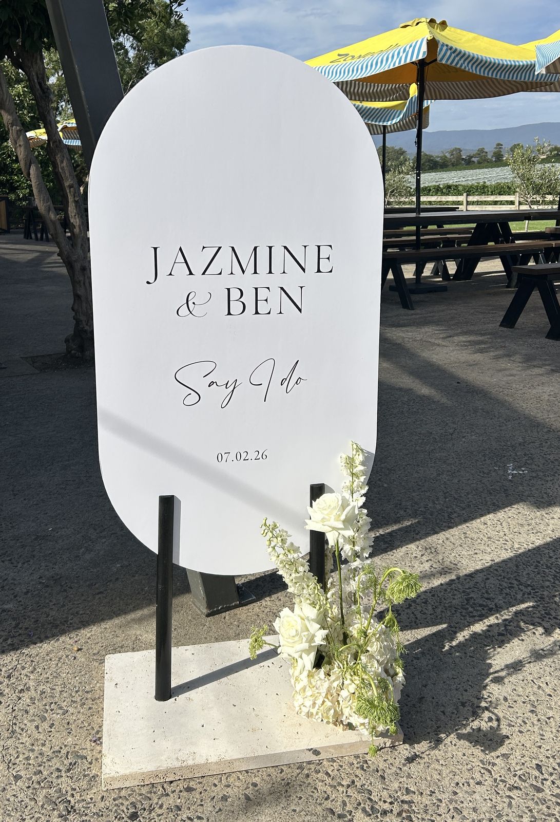 White pill-shaped welcome sign reading Jazmine and Ben, Say I do, on a slim black metal stand with a white base, outdoors