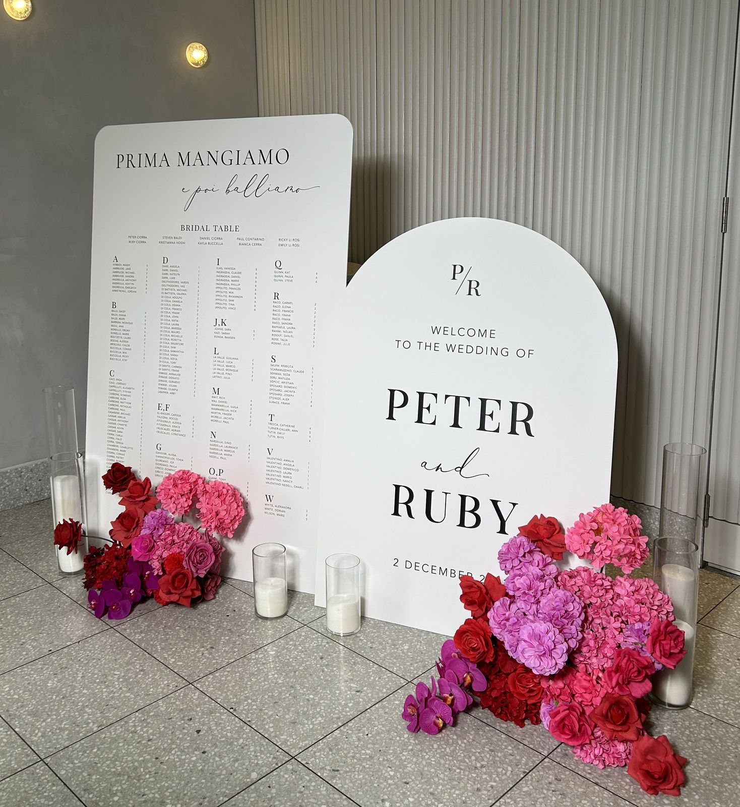 White rounded seating chart headed Prima mangiamo e poi balliamo with a bridal table section and alphabetical columns, beside a white arch welcome sign for Peter and Ruby with pink and red florals