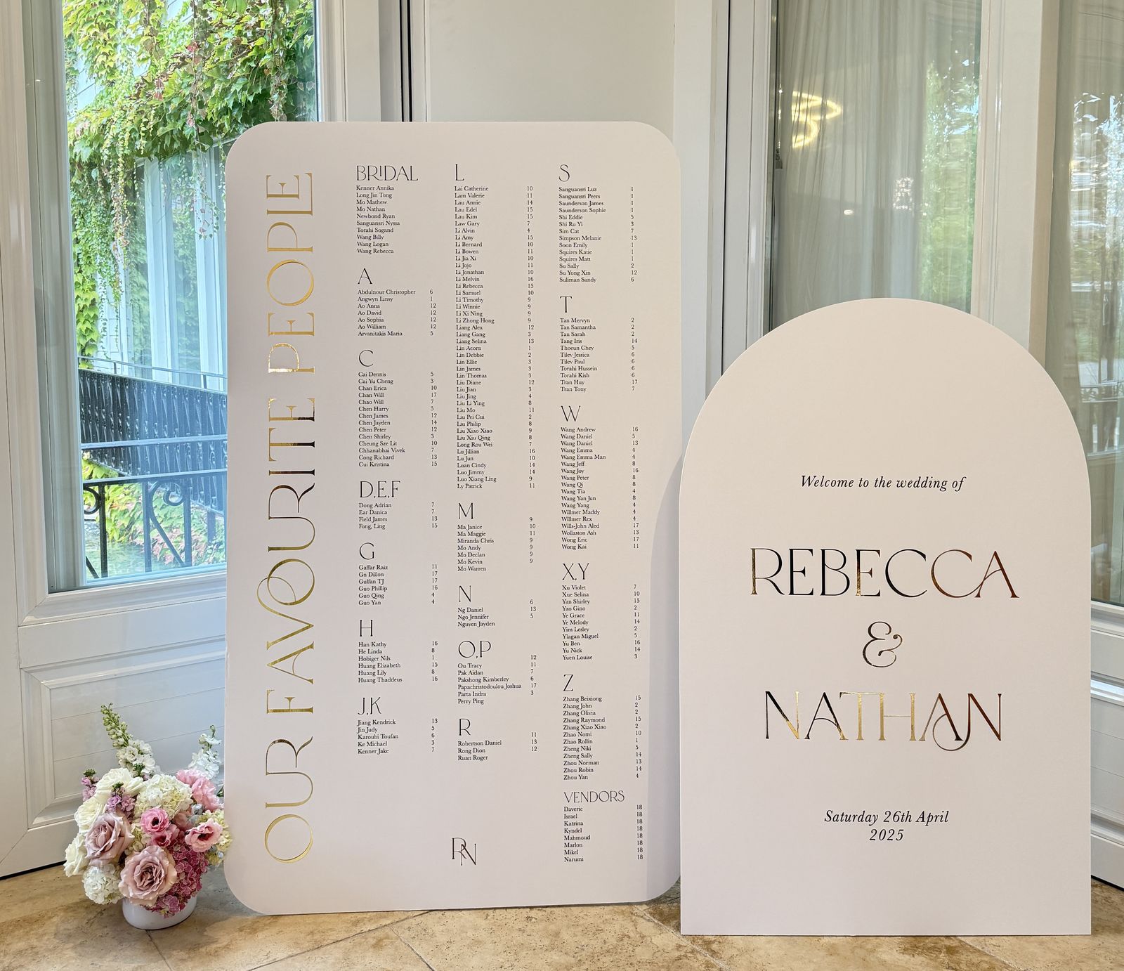 White rounded seating chart with Our favourite people in gold mirror lettering running vertically, alphabetical columns and a vendors section, beside a white welcome sign for Rebecca and Nathan
