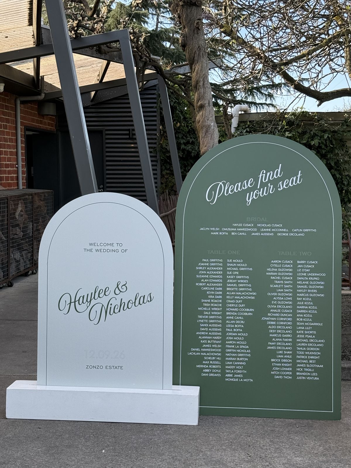 Deep green arch seating chart reading Please find your seat with guests listed by table, beside a pale blue arch welcome sign on a white plinth