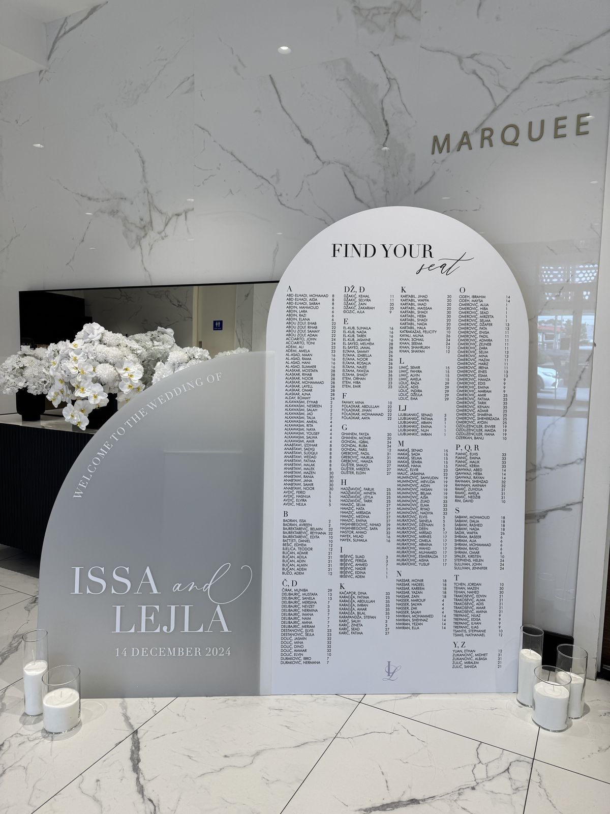 Grey half-arch welcome sign with Welcome to the wedding of set along the curved edge, the names Issa and Lejla below, beside a white arch Find your seat chart