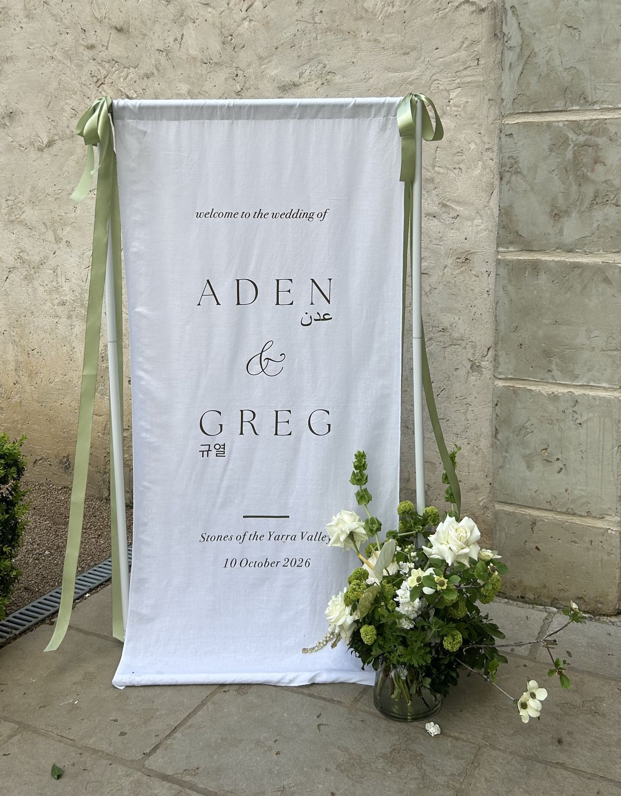 White linen welcome sign for Aden and Greg with their names also written in Arabic and Korean script, hung from a white stand with sage green ribbons beside a vase of white roses