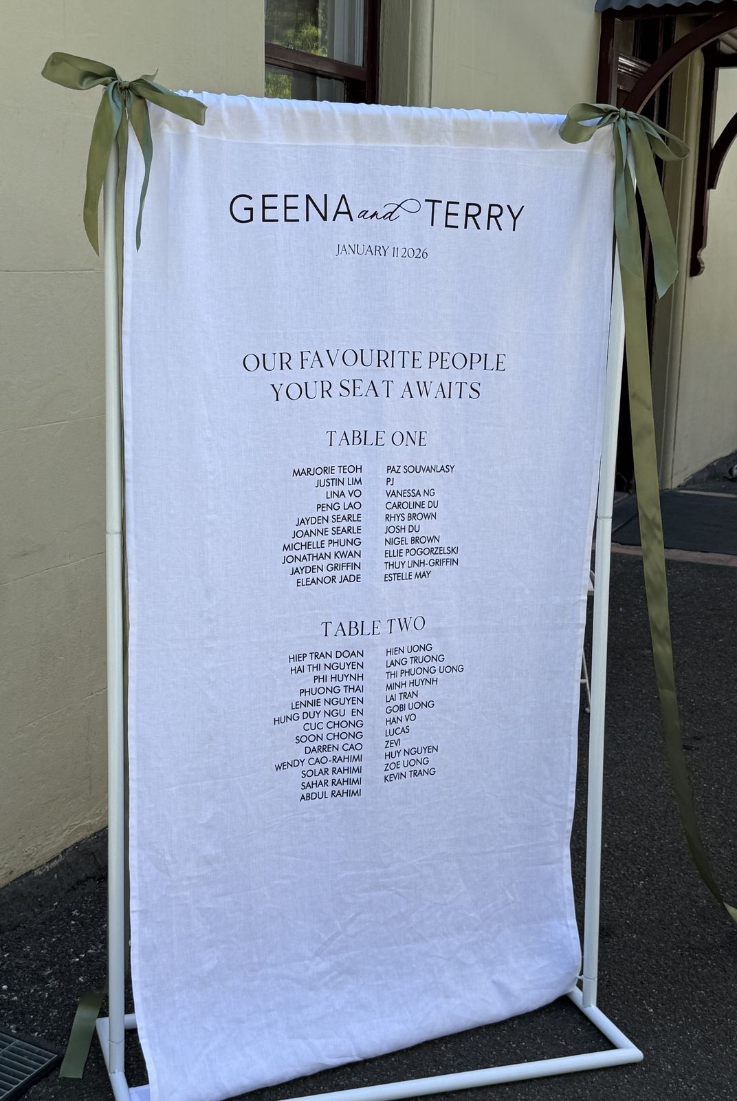White linen seating chart tied to a white frame with sage ribbons, showing the couple's names and date, then guests listed under Table One and Table Two