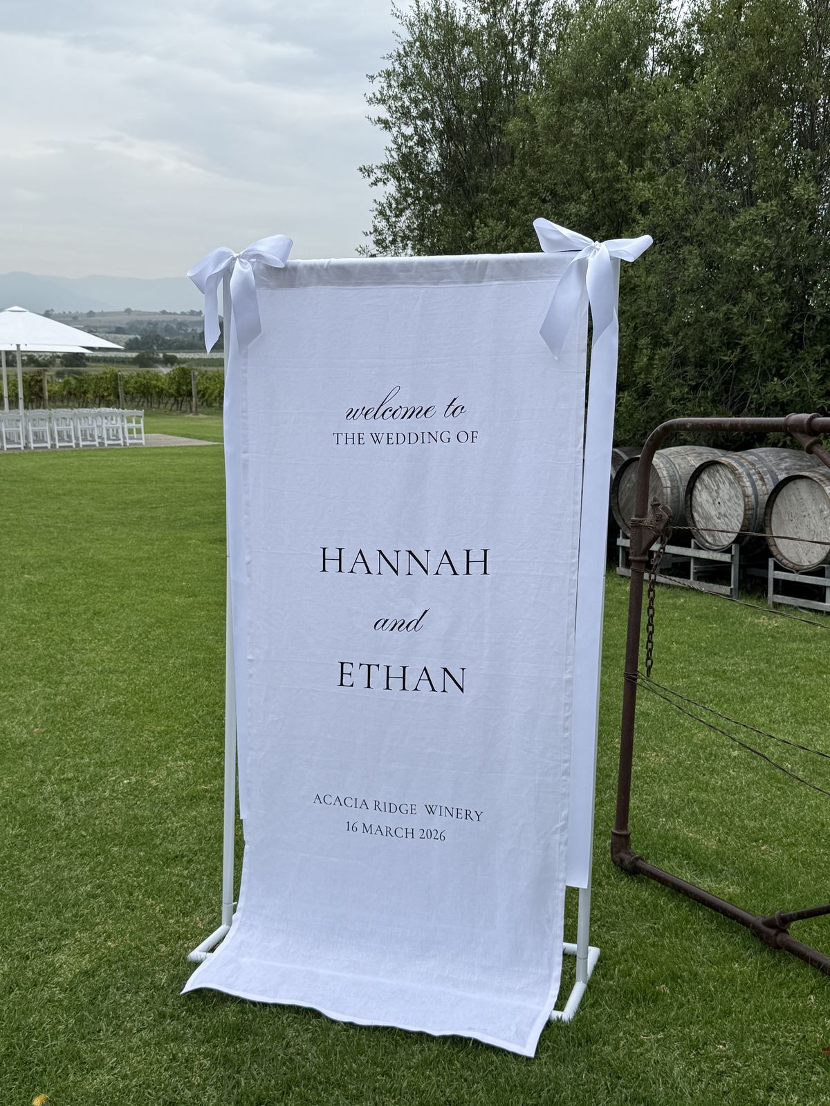 White linen welcome sign tied to a white frame with white bows, reading welcome to the wedding of Hannah and Ethan, Acacia Ridge Winery, standing on a lawn