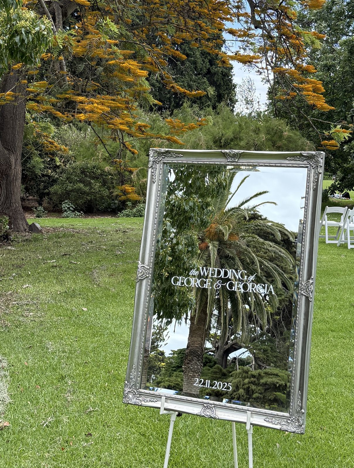 Silver mirror wedding welcome sign in an ornate silver frame on a white easel in a garden, reflecting a palm tree