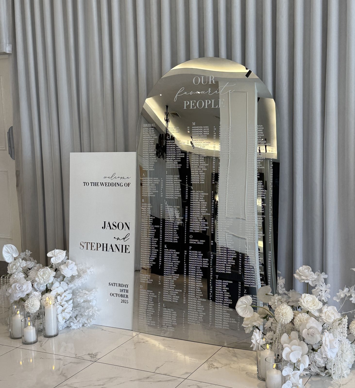 Silver mirror arch seating chart headed Our favourite people, with a white welcome sign for Jason and Stephanie in front and white florals and candles at the base