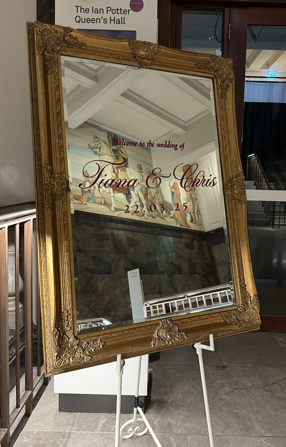 Ornate gold-framed mirror wedding welcome sign with burgundy script lettering for Tiana and Chris on a white easel