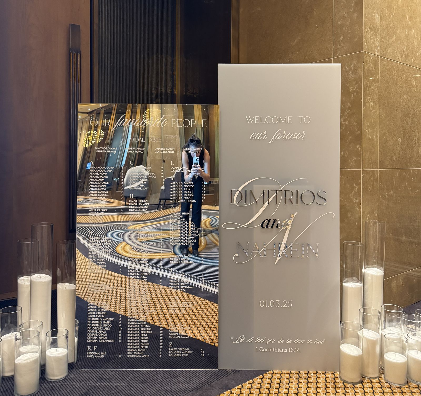 Mirror seating chart reflecting a gold patterned carpet and a guest taking a photo, beside a frosted welcome sign with a monogram, surrounded by pillar candles