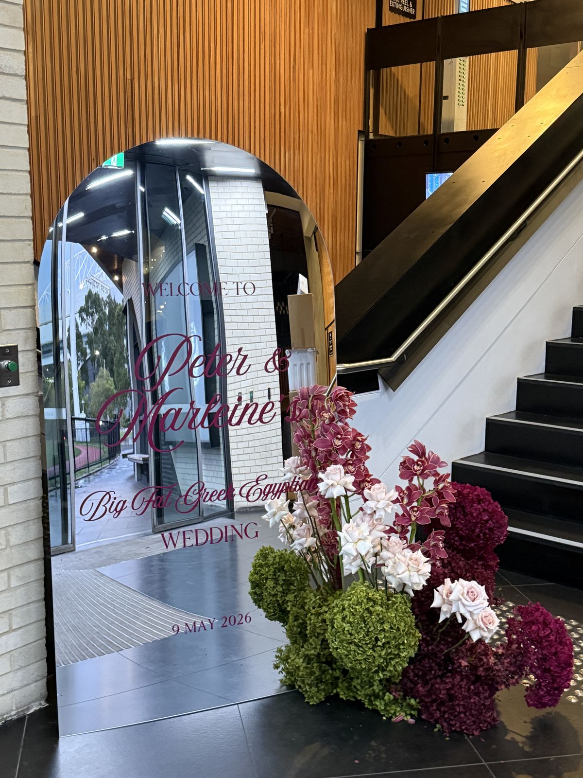 Silver mirror arch welcome sign with plum lettering for Peter and Marleine, with burgundy, green and pink florals at the base beside a staircase