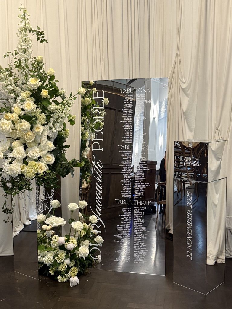Tall mirror panel seating chart listing guests under Table One, Table Two and Table Three, beside a mirror plinth with a vertical date, surrounded by white roses