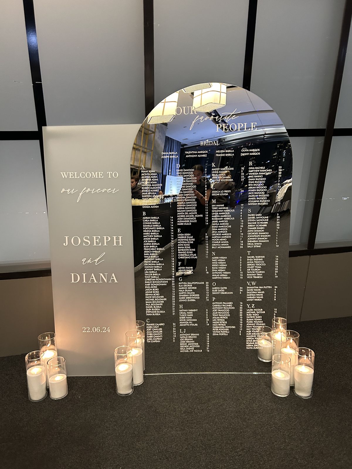 Silver mirror arch seating chart headed Our favourite people listing guests alphabetically with a table number beside each name, next to a frosted acrylic welcome sign for Joseph and Diana, surrounded by candles