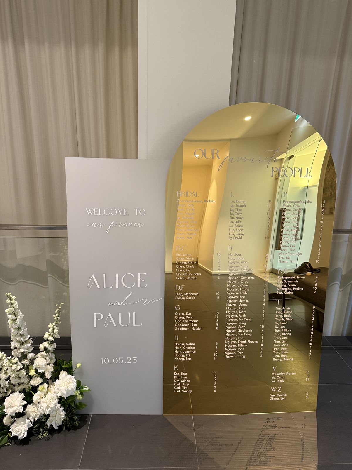 Frosted acrylic welcome sign reading Welcome to our forever, Alice and Paul, in front of a gold mirror arch seating chart with white flowers at the base
