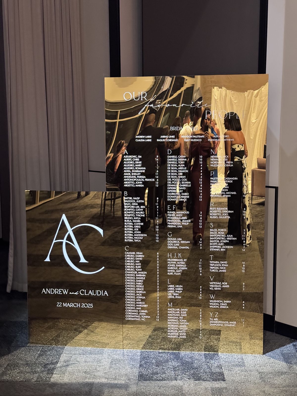 Two overlapping gold mirror panels: the lower one with a large AC monogram and the names Andrew and Claudia, the upper one with an alphabetical seating chart