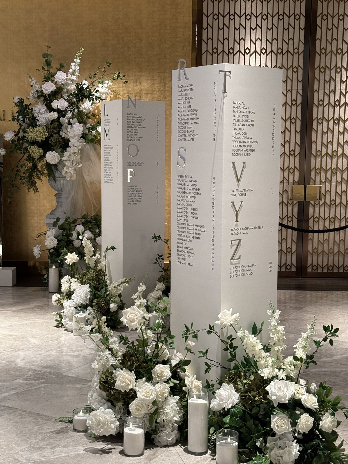 Tall white pillars printed with seating chart names grouped by large letters such as R, S, T, V, Y and Z, surrounded by white roses and candles