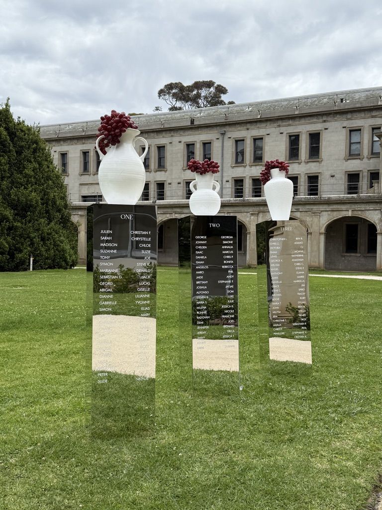 Row of mirror plinths on a lawn, each listing the guests for one table such as One and Two, topped with white vases holding red grapes, in front of a heritage building