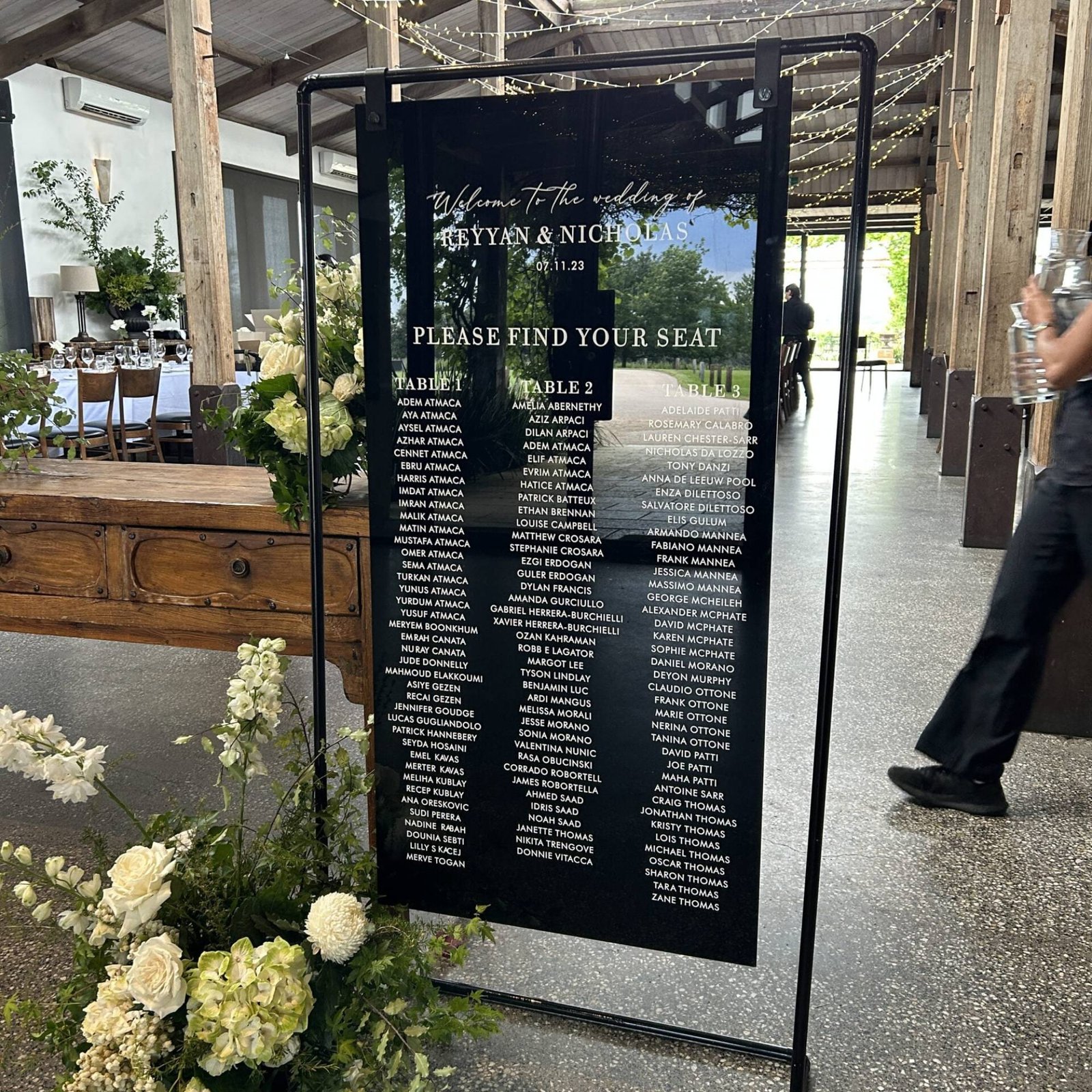 Black acrylic sign in a black metal frame stand reading Welcome to the wedding of Reyyan and Nicholas, Please find your seat, with guests listed under Table 1, Table 2 and Table 3