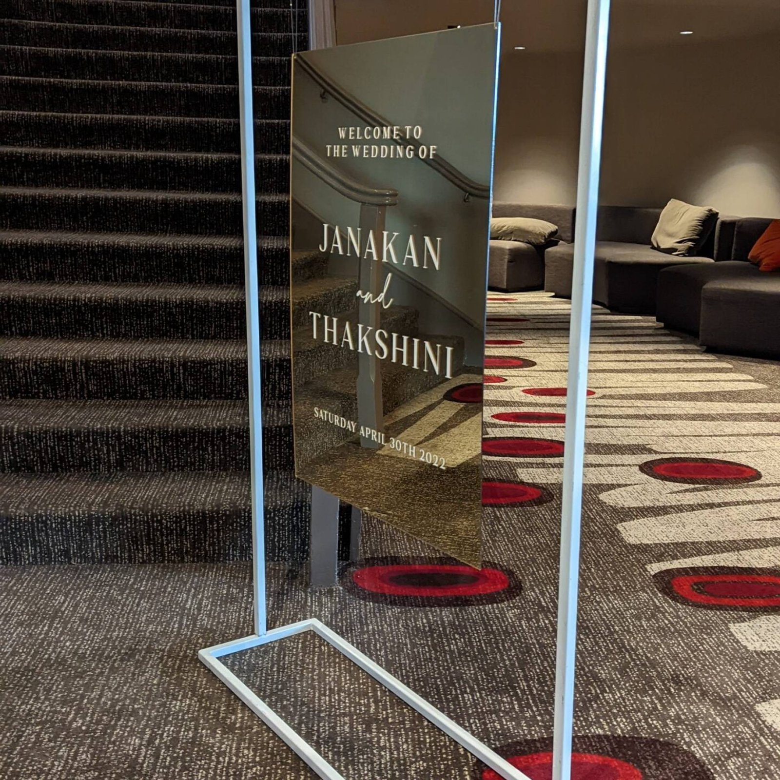 Gold mirror welcome sign for Janakan and Thakshini hanging in a white metal frame stand beside a carpeted staircase