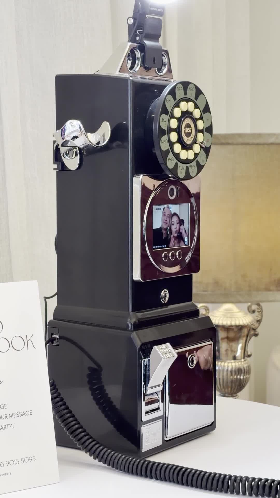 Black retro payphone-style video guest book with a chrome dial and a built-in screen showing two guests recording a message